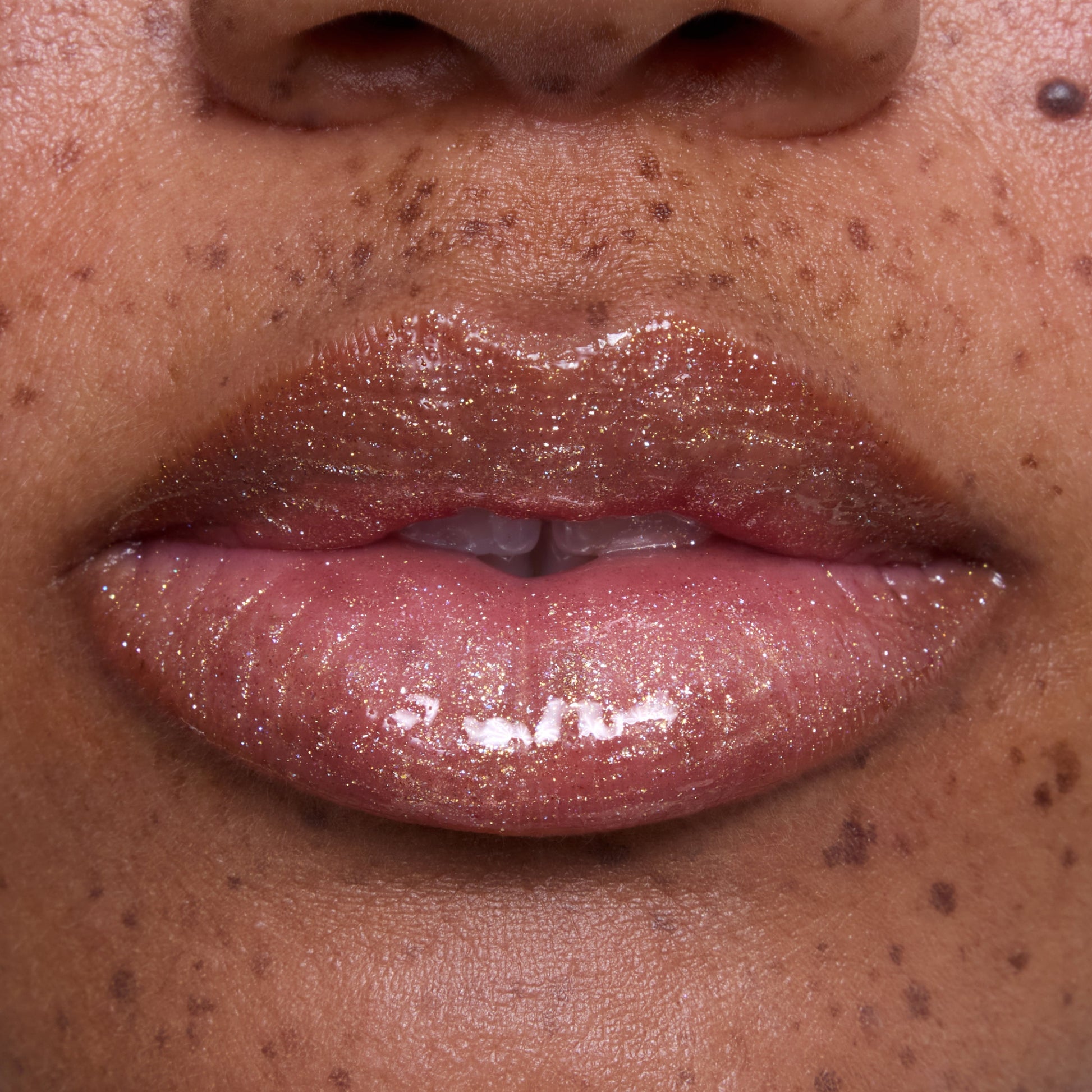 Close-up of glossy, medium-toned lips wearing Ambitious Tiana Ultra Glossy Lip, showing shimmering gold and pink sparkle inspired by Disney Princess beauty, with natural skin and freckles softly visible around the mouth.