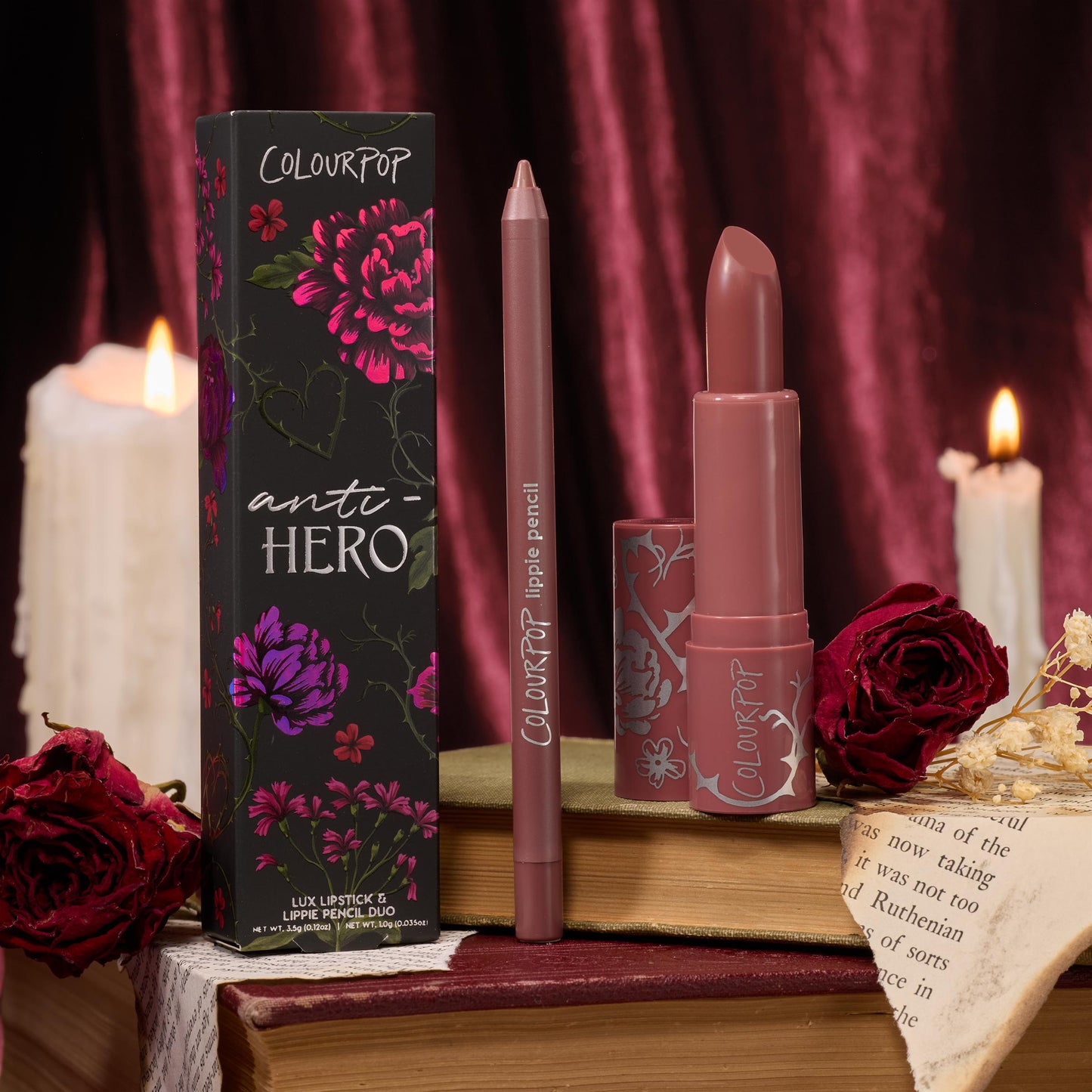 Anti-Hero ColourPop Lux Lipstick and matching lippie pencil stand uncapped beside the floral box, revealing a rosy-mauve bullet; displayed on vintage books with red roses and dripping candles against burgundy velvet for a moody, romantic vibe.